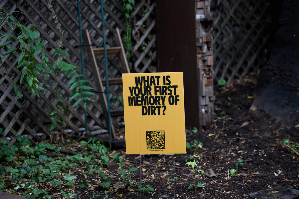"What is your first memory of dirt?" Black text on a yellow background accompanied by a QR code. This image shows the graphic on a lawn sign in a green backyard space.