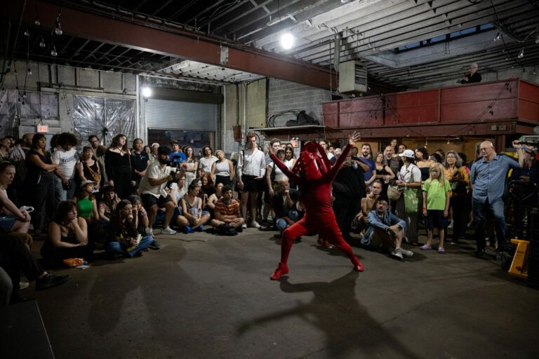 A crowd gathers around a person wearing a red costume. They all watch from inside an industrial building.