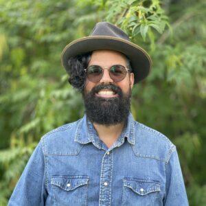 A photo of artist Osvaldo Budet in front of foliage. He wears a hat and sunglasses and grins at the camera.