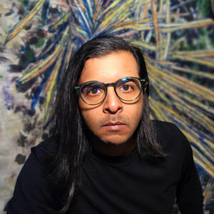 A close up photo of artist Pseudomero. He wears glasses and looks intently at the camera.