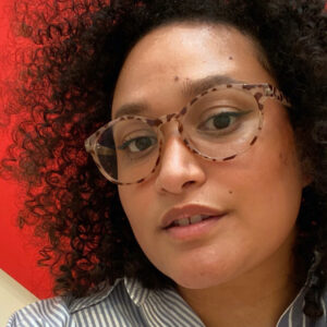 A close up photo of artist Estefanía Vallejo Santiago, also known as "Saint Electric." She wears glasses and smiles lightly at the camera.