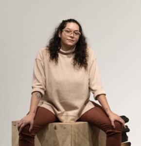 A seated photo of artist Valeria Nicolás Tizol Vivas. She wears glasses and smiles lightly at the camera.