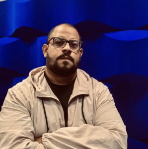 Artist Jason "Baco" Ortiz stands, arms crossed, in front of a wavy blue wall. He wears glasses and looks at the camera with a neutral expression.