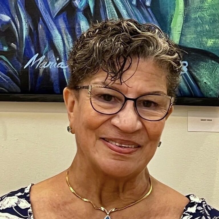 A close up photo of artist Maria Dominguez standing in front of her artwork. She wears glasses and smiles at the camera.