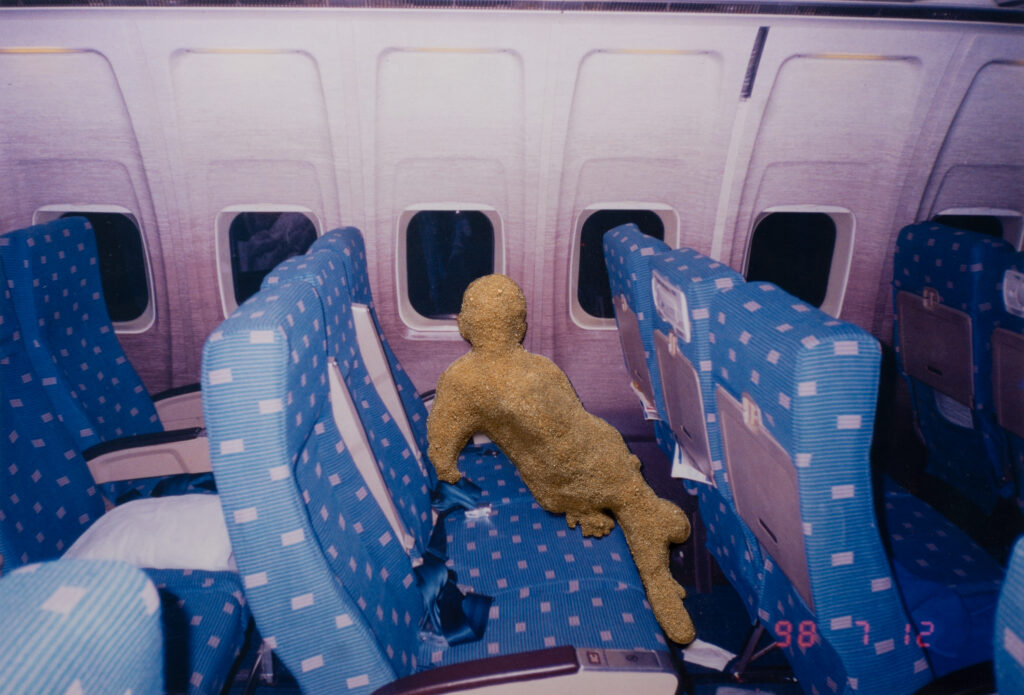 An image of the interior of a plane. A sand pile in the shape of a child sits in one of the plane chairs.