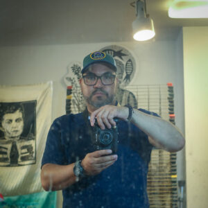 A self portrait of artist Oliver Rios. He holds his camera in front of his chest and looks at the viewer with a neutral expression.