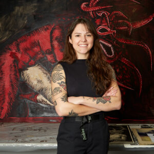A headshot of artist Elisa Dore in her studio. She stands in front of her artwork.