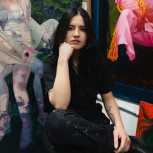 Artist Isabella Mellado sits in front of pieces of their artwork. Her head is propped up on her hand and she looks at the camera with a neutral expression.