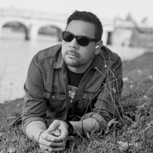 A grayscale photo of artist Rafael Miranda Mattei laying on a grassy hill and propped up on his arms. He is wearing sunglasses and looking away from the camera with a neutral expression.