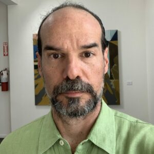 A close up photo of artist Miguel Trelles. He stands in front of a painting and looks at the camera with a neutral expression.