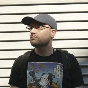 A photo of artist David Arroyo. He wears glasses and looks away from the camera with a neutral expression.