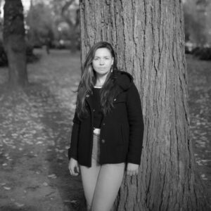 A grayscale headshot of artist Gabriella N. Báez standing in front of a large tree. They look at the camera with a neutral expression.