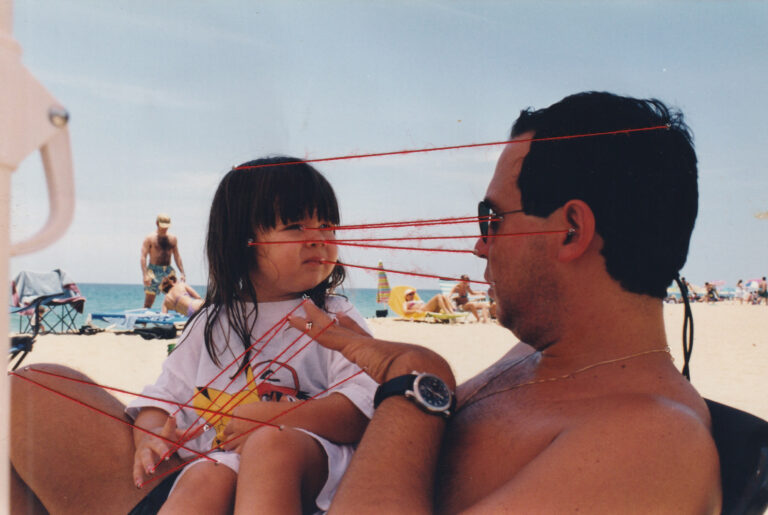 A photograph of a young person sitting on their father's lap at the beach. There are red embroidered threads connecting their fingertips, knees, eyes, ears, backs of the head, noses, and mouths.