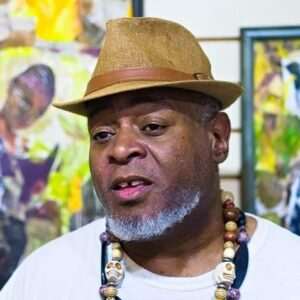 A close up photo of artist Ram Brisueño standing in front of his artwork. He wears a hat and looks off camera with a neutral expression.