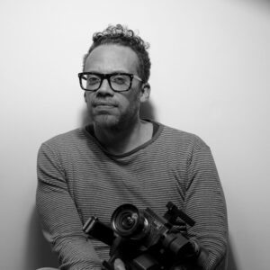 A grayscale photo of artist Carlos Javier Ortiz holding a camera. He wears glasses and looks at the camera with a neutral expression.