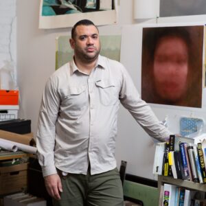 Artist Dennis Delgado stands in his studio, facing the camera with a neutral expression. Behind him are multiple pieces of his artwork.