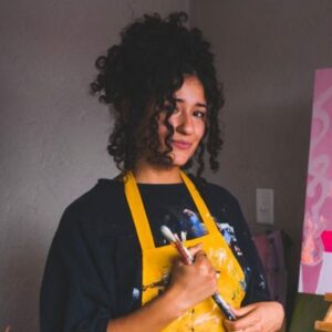 Artist Emily Rose stands smiling in her studio holding paint brushes Behind her is a piece of her artwork.