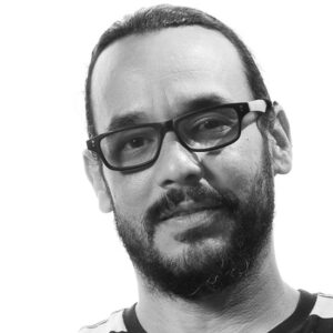 A grayscale close up photo of artist Josué Guarionex. He wears glasses and smiles lightly at the camera.