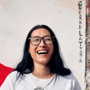 Artist One Rad Latina stands in front of their artwork. She wears glasses and laughs facing the camera.