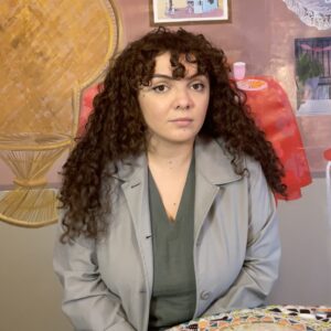 A photo of artist Misla seated in front of her artwork. She looks at the camera with a neutral, open expression.