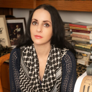 A photo of artist Molly Crabapple. She wears and color inverted keffiyeh and looks at the camera with a neutral, open expression.