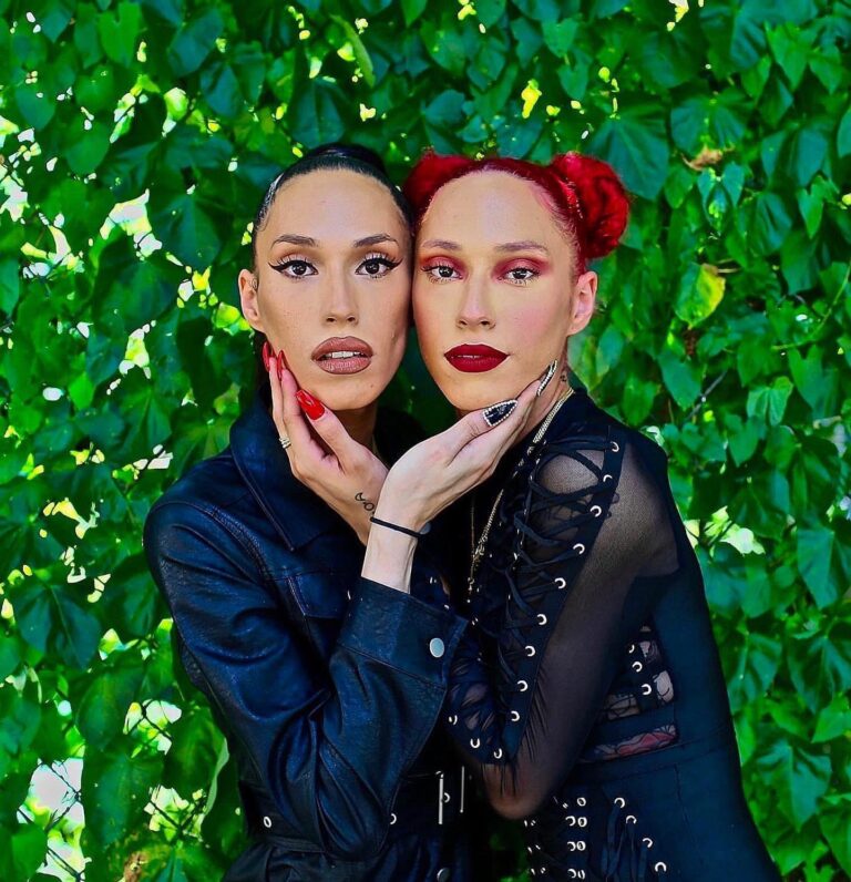 Twins Kykki and Ari stand with their bodies facing each other but their faces towards the camera. They caress each other's face with one hand. The background is full of leaves.