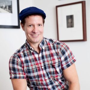 A seated headshot of artist Ruben Natal-San Miguel. He wears a blue hat and grins at the camera.