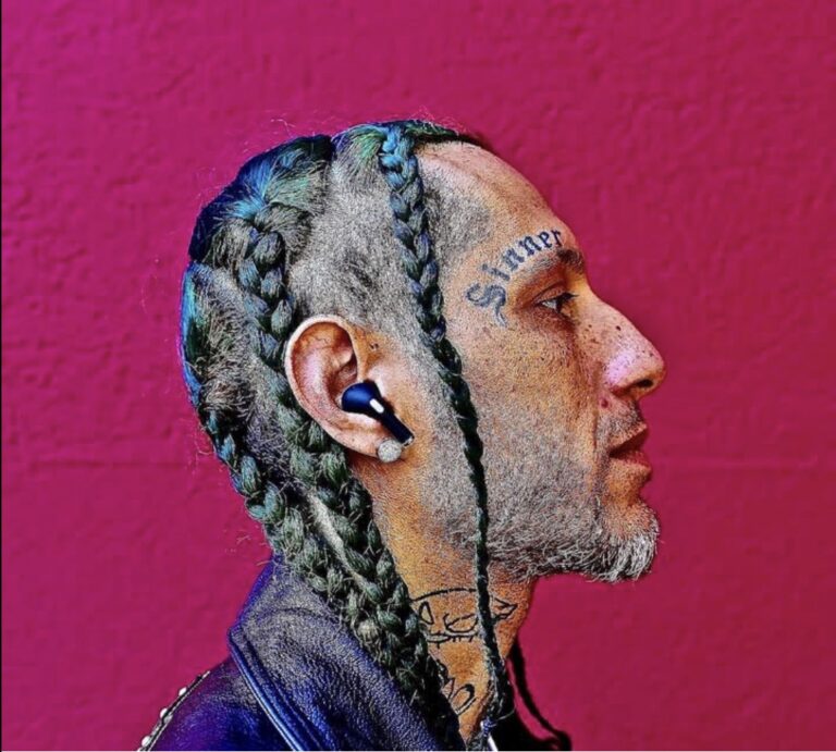 A man with braids in his hair faces right. Tattooed above his eyebrow is the word "Sinner." He also has tattoos on his neck. He wears a Bluetooth earbud.