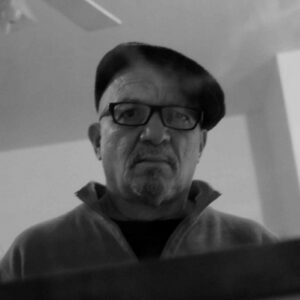 A close up grayscale photo of Máximo Rafael Colón. He wears glasses and a hat and looks above the camera with a neutral expression.
