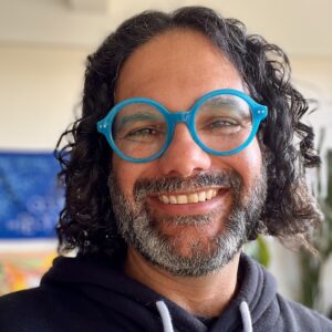 A close up photo of artist Hatuey Ramos-Fermín. He wears bright blue glasses and grins at the camera.