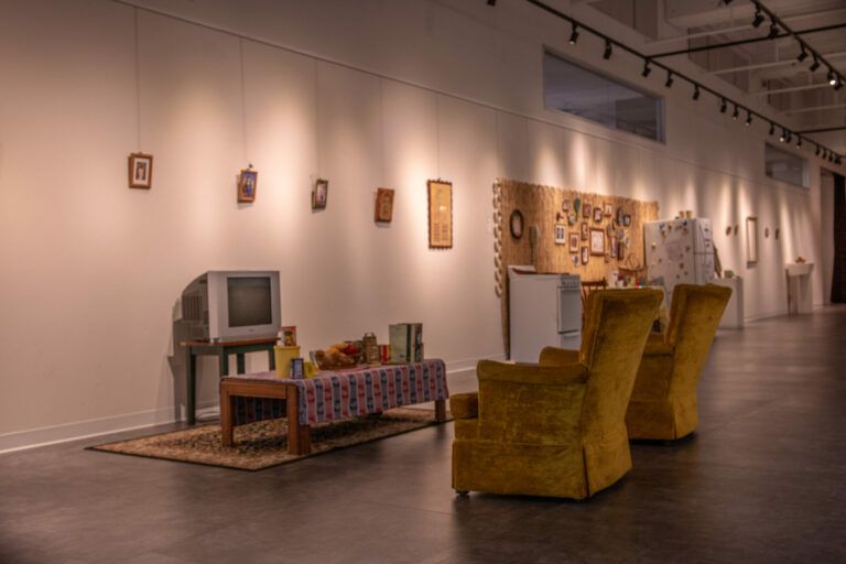 A living room of two chairs, a table, and a tv with family pictures hanging from the wall. Behind the living room setup there is a kitchen set.