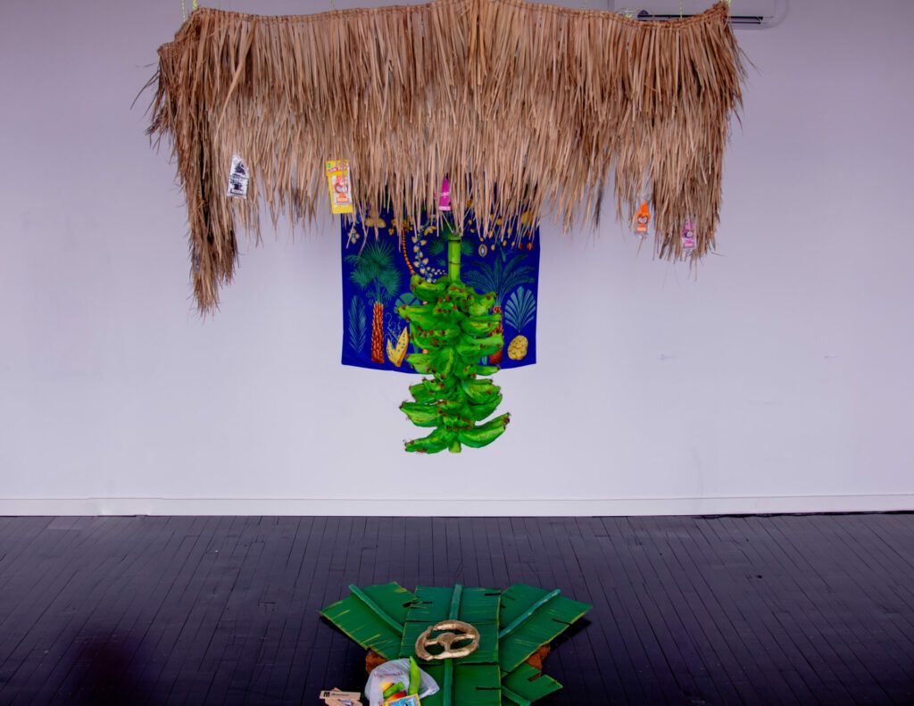 Dried palm with attached car air-freshener trees hanging over a branch of bananas/platanos. Under the overhang there is a golden steering wheel, groceries, and dominos. On the wall behind it all there is a bold tapestry of trees.