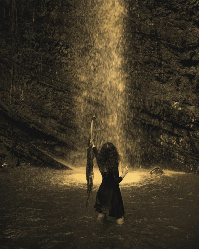 A golden-hued image of a woman standing thigh-deep in water under a waterfall. she holds up a palm frond to the falling water away from the camera.