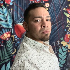 A close up of artist Jean Carlos Valentin Velilla. They stand in front of a floral backdrop facing away. His head is turned to look at the camera with a neutral expression.