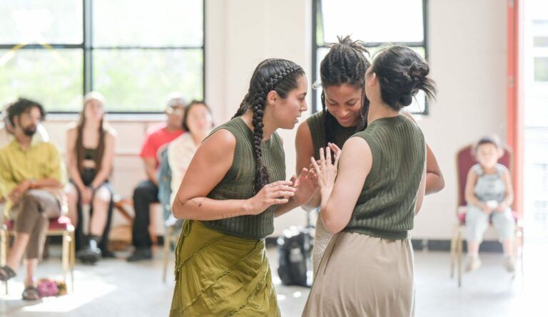 Brianna Torres, Chinchin Hsu, Natalya Shoaf and Bria Monet Barfield are standing in a circle with their arms reaching out to each other's backs. They are in a well-lit room with other people sitting behind them in the distance.