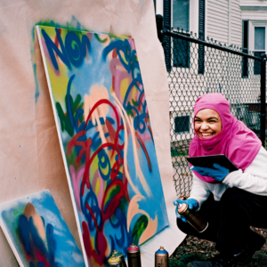 A photo of artist Victoria "Thirteenvic" DelValle at work. She has turned away from her piece and is laughing at something off camera.
