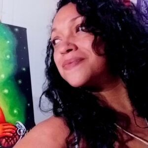 A close up photo of artist Yasmín Hernández. A piece of her artwork is partly in frame beside her. She faces away from the camera and smiles.