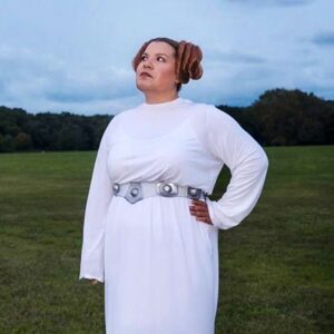 A photo of artist Rhynna M. Santos in a field dressed as Princess Leia from Starwars. She looks away from the camera with a neutral expression.