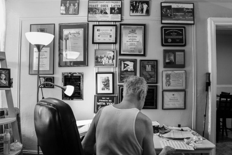 The desk of the artist's father and music legend Ray Santos. The wall above his desk is covered in awards and news clippings. Ray Santos is studying sheet music at his desk.