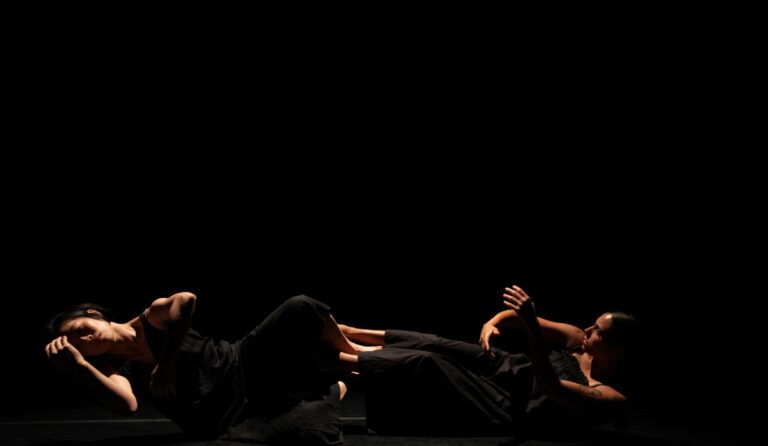 Brianna Torres and Chinchin Hsu dance as they lay down on their sides. They both wear full black costumes and the background is full black.