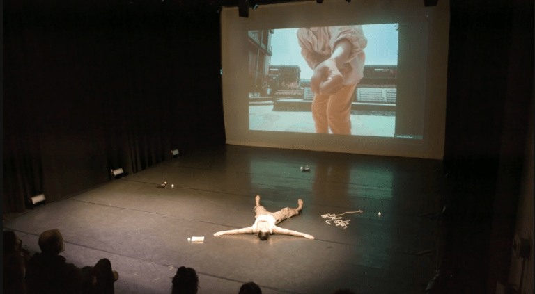 Still from the 'True North" performance. A spotlight illuminates a dancer sprawled out on their back on a dark stage. There is a screen on the back of the stage that currently shows a person holding out their fist to the camera.