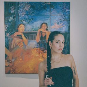 A photo of artist Beatriz Whitehill. She stands in front of her artwork with a neutral expression.