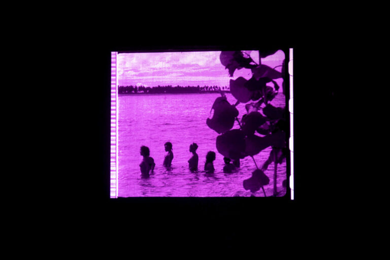 Partially nude women stand waist-deep in the ocean, seen through some round leaves. The image is a bright magenta.