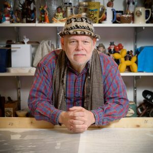 A photo of artist Pablo Delano seated in front of crowded shelves. He wears a hat and looks into the camera with a neutral expression.