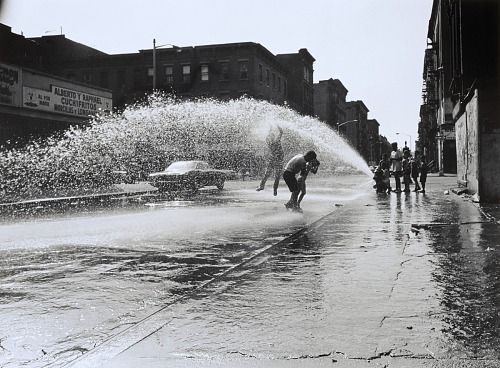 Hydrant water sprays in a high arc over a few people in a NYC street. They shield their faces and jump to touch the water. A few other figures stand off behind the hydrant.