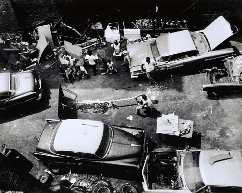 Bird's-eye-view of people gathered in an alley surrounded by cars. A central figure roasts a pig on a spit over a fire connected to a custom rig.