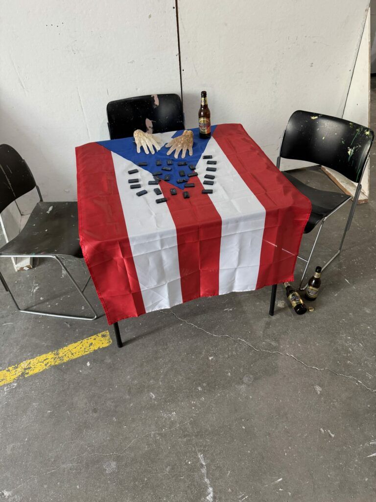 A table draped with a Puerto Rican flag and covered in black domino-shaped objects. There are resin hands at the table next to a Medalla Light bottle.