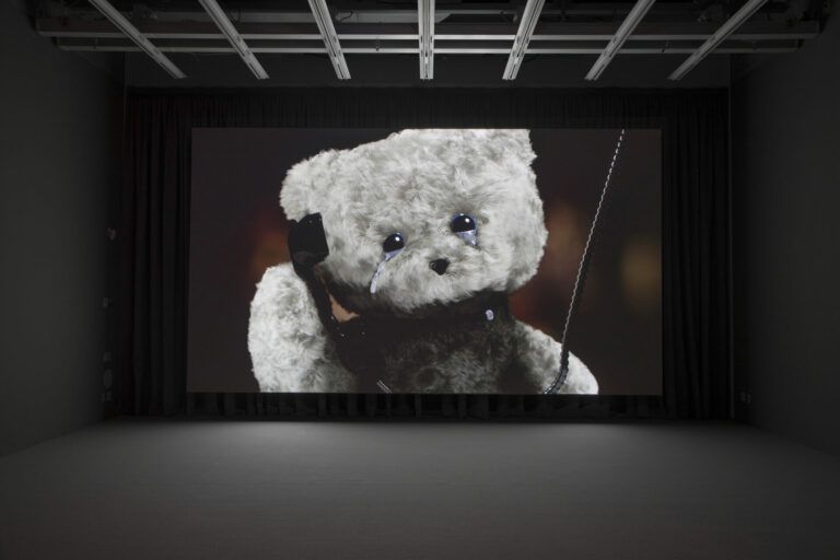 Still from the 3D animated film, "Moléculas." It shows a grey bear with a black torso from the chest up. The bear holds a payphone up to its ear while crying.