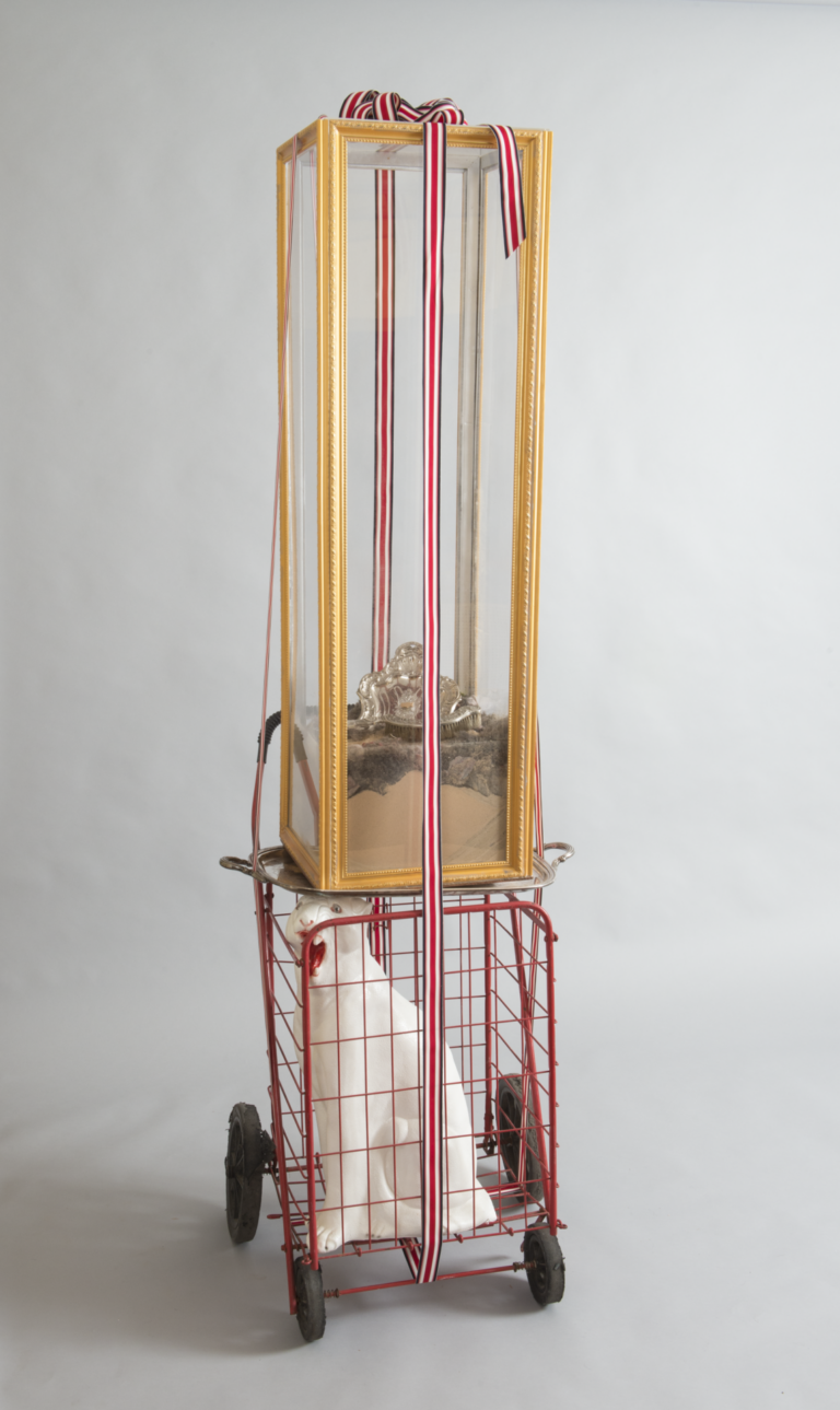 Tall glass box with gold trim containing dust from Puerto Rico's capital and a dustpan. The box is strapped to a red cart with a white cat figure inside.
