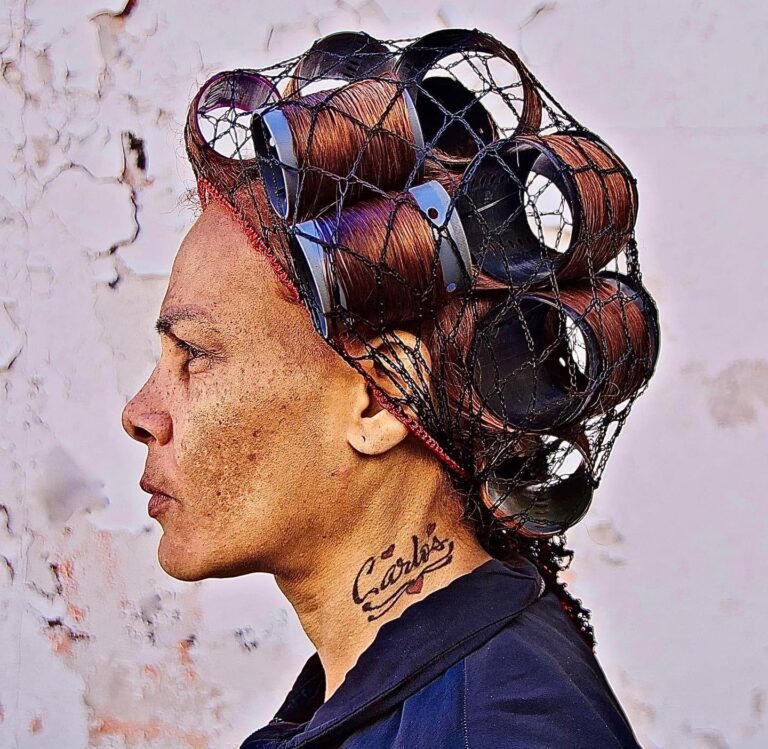 A woman facing to the left displaying the tattoo "Carlos" on her neck surrounded by hearts. She is wearing hair curlers and a hairnet.